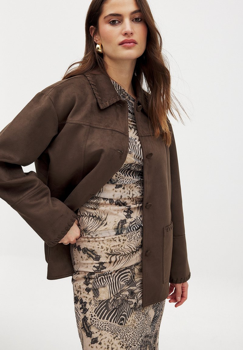 Brown faux suede jacket with a collar, button closure, and side pockets, layered over a patterned dress featuring animal prints and textures.