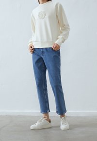 Sudadera crema con un diseño floral texturizado en el pecho, combinada con jeans de cintura alta azul y zapatillas blancas con un acento en color beige.