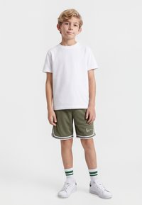Niño de pie con camiseta blanca, pantalones cortos deportivos verde oliva con rayas blancas, zapatillas blancas y calcetines blancos con rayas verdes.