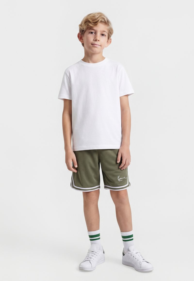 Niño de pie con camiseta blanca, pantalones cortos deportivos verde oliva con rayas blancas, zapatillas blancas y calcetines blancos con rayas verdes.