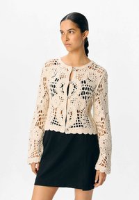 Virkat cardigan i krämfärg med blommönster, öppen väv, knäppning och långa ärmar, bärs över en svart klänning.