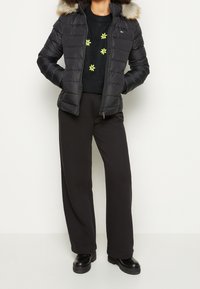 Chaqueta acolchada negra con capucha forrada de piel, cierre de cremallera y costuras horizontales. Lleva un suéter negro con bordado floral amarillo debajo.