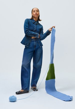 Veste en denim et jean à jambes larges en délavage bleu moyen, associés à des chaussures plates noires. Une longue pièce en crochet rayé présente des segments bleus et verts.