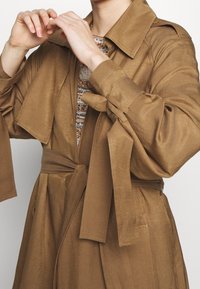 Beżowy płaszcz trench z paskiem, szerokimi rękawami i kołnierzem. Materiał ma gładką fakturę. Widać warstwową bluzkę w wzory spod płaszcza.