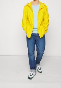 Person med gul regnjakke, lyseblå skjorte, optrullede blå jeans og hvide sneakers med grønne accenter, stående mod en ensfarvet baggrund.