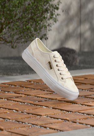 Białe niskie buty sportowe z beżowymi wzorami, złotym logo „GUESS” oraz białą gumową podeszwą, umieszczone na drewnianej powierzchni z przerwami wypełnionymi żwirem.