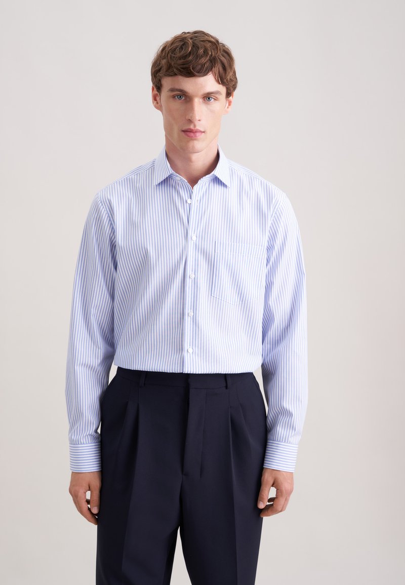Homme portant une chemise boutonnée bleu et blanc à rayures verticales, rentrée dans un pantalon noir plissé taille haute, debout devant un fond uni.