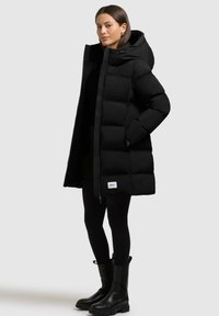Cappotto imbottito nero con cappuccio, caratterizzato da un design trapuntato, zip frontale e tasche laterali. Abbinato a leggings neri e stivaletti.