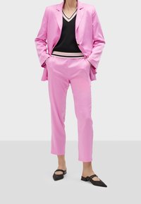 Personne portant un costume rose vif avec un blazer cintré et un pantalon court, un top noir en V et des ballerines noires pointues, debout les mains dans les poches.