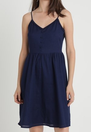 Kobieta w granatowej, bezrękawnikowej sukience z cienkimi ramiączkami, dekoltem w serek i guzikami na przodzie na jednolitym tle.