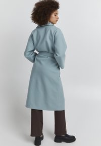 Manteau bleu clair ceinturé avec un large col, des manches longues et une texture lisse, associé à un pantalon large marron et des bottes noires imposantes.