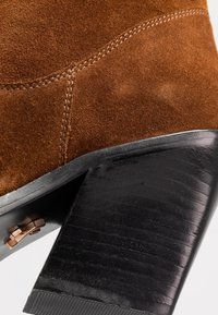 Brauner Wildleder-Stiefelette mit schwarzem, klobigem Absatz. Verfügt über abgestepptes Detail und ein Metallakzent in der Nähe des Absatzes. Glatte Textur.