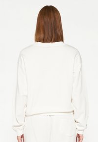 Sudadera blanca de gran tamaño con mangas largas, puños ajustados, cuello redondo y textura similar al ante. Cuenta con un detalle de bolsillo trasero.