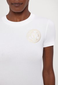 Bijela uska majica s kratkim rukavima izrađena od pamuka, s zlatnim vezom u obliku kruga i natpisom "Versace Jeans Couture" na lijevoj strani prsnog područja.