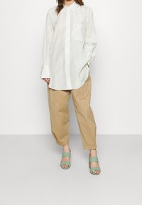 Chemise blanche à manches longues avec un motif subtil, associée à un pantalon beige clair ample et des sandales à talons tissées vertes.