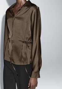 Brown satin shirt with button front, long sleeves, and drawstring waist. Smooth texture and tailored fit, paired with black trousers.