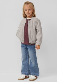 Graue Bomberjacke mit Reißverschluss, bordeauxrotes Hemd darunter, bell-bottom Jeans in Blau und weiße Sneakers mit braunen Akzenten. Mädchen mit langen blonden Haaren.
