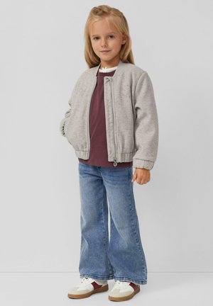 Grijze bomberjack met een rits, bordeaux shirt eronder, uitlopende blauwe jeans en witte sneakers met bruine accenten. Meisje met lang blond haar.