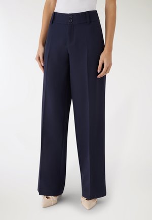 Persona che indossa pantaloni navy a vita alta con gamba larga, chiusura con doppio bottone e tacchi beige con punta appuntita, in piedi su sfondo bianco.