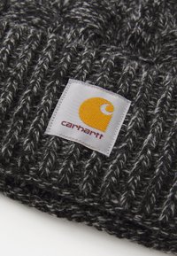 Gorro de punto gris con un patrón texturizado. Incluye una etiqueta blanca cuadrada con un logotipo amarillo y la palabra "carhartt" en texto burdeos.