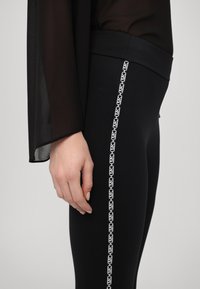 Czarne legginsy o gładkiej fakturze mają białą wzorzystą boczną lamówkę. Powyżej częściowo widoczna jest przezroczysta czarna bluzka z rozszerzanymi rękawami.