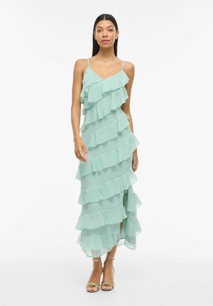 Naine, kes kannab helerohelist, õhukesi rihmasid ja kihilist volangidega varrukateta kleiti ning kuldseid kõrge kontsaga sandaale, seisab valge tausta ees.