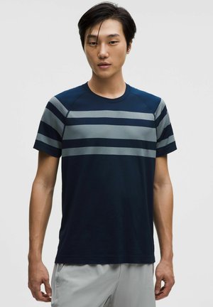 Junger Mann trägt ein marineblaues Kurzarmhemd mit grauen horizontalen Streifen, steht vor einem schlichten hellen Hintergrund.