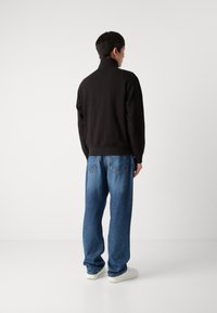 Felpa nera con collo alto, dal tessuto morbido, abbinata a jeans denim blu a vestibilità ampia e sneakers bianche, vista da dietro.