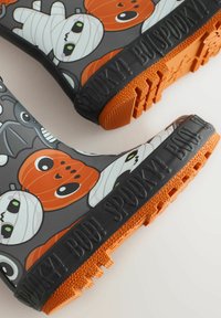 Bottes en caoutchouc de pluie sur fond gris, ornées de citrouilles, de momies et de chauves-souris en dessin animé. La semelle est orange avec une texture antidérapante.