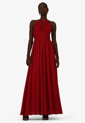 Frau trägt ein bodenlanges, ärmelloses rotes Abendkleid mit gerafftem Oberteil und Neckholder-Ausschnitt, steht mit entspannten Armen an den Seiten.