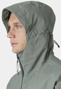 Hedvábná bunda v tlumené zelené barvě, vyrobená z lehkého materiálu. Obsahuje zipový uzávěr, kapuci se šňůrkou a viditelné švy.
