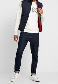 Homme portant un gilet matelassé bleu marine avec doublure rouge par-dessus un sweat-shirt blanc avec l'imprimé "69", un jean bleu foncé avec revers, et des baskets blanches.