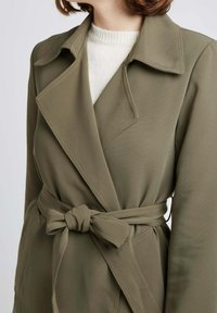Trench-coat vert olive avec un design enroulé, un col cranté et une ceinture nouée à la taille. Tissu lisse avec des coutures visibles.