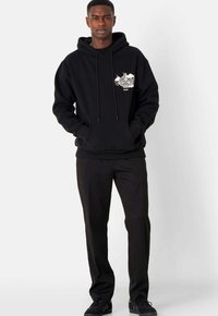 Sudadera negra con un bolsillo frontal, cordones y un diseño gráfico en blanco en el lado izquierdo del pecho. Combinada con pantalones y zapatos negros.