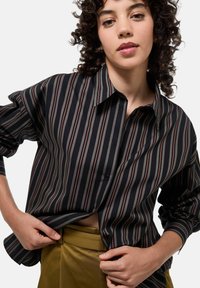 Chemise à rayures noir et blanc avec des accents rouges et verts, design à boutons, manches longues, coupe légèrement ample, associée à un pantalon vert olive.