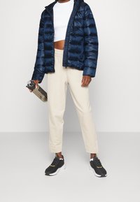 Person som bär en marinblå puffjacka över en vit kort ovan och beige byxor, håller en genomskinlig vattenflaska, står på en grått golv.