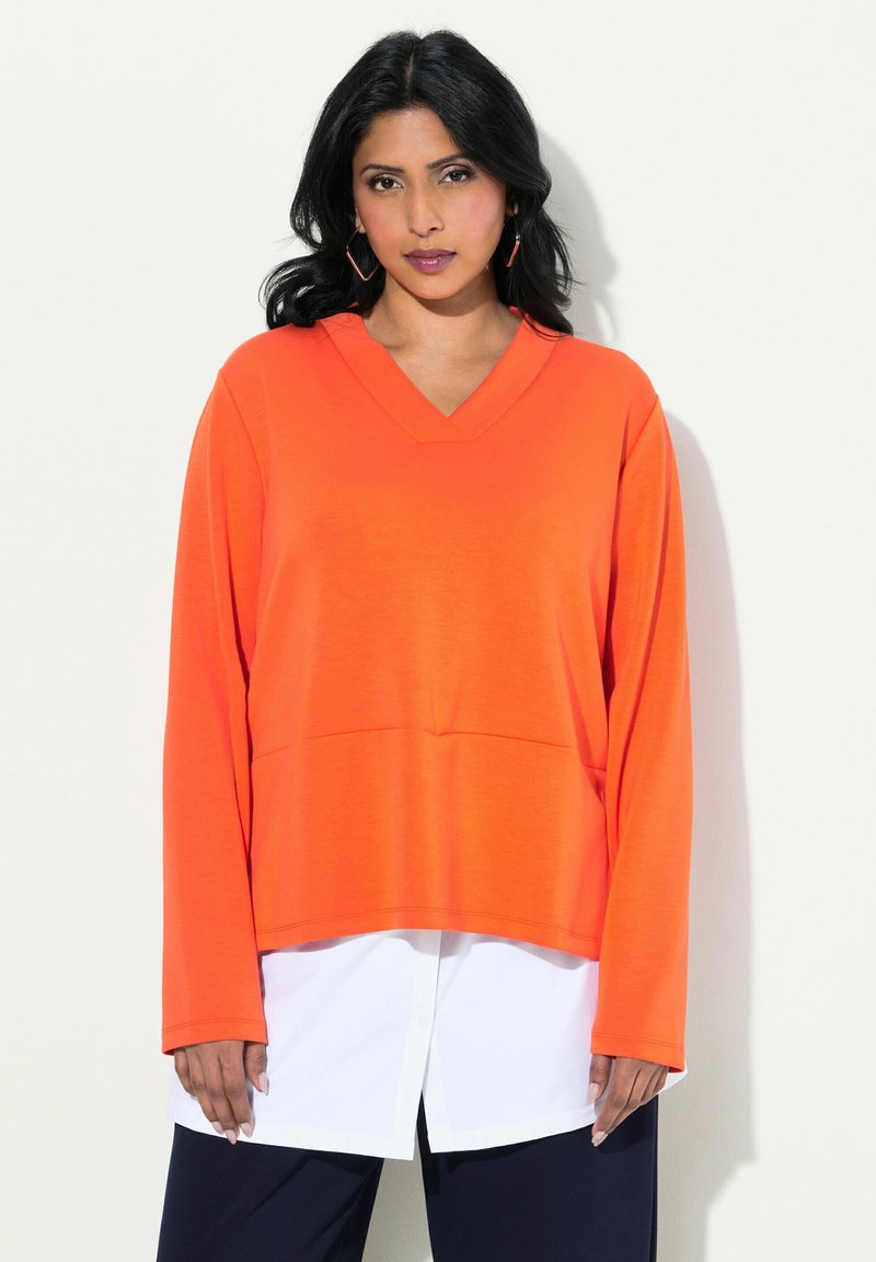 Maglietta a maniche lunghe con scollo a V in tessuto arancione brillante con una vestibilità comoda, sovrapposta a una camicia bianca. Dettaglio della tasca frontale piatta.