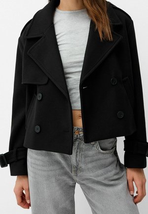 Kvinde iført en kort sort dobbeltradet jakke over en lysegrå crop top og falmede grå jeans, stående med afslappede arme.
