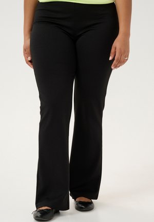 Pantalons noirs extensibles avec un design à jambe évasée, fabriqués en tissu doux. Associés à des ballerines noires, avec un simple nœud décoratif.
