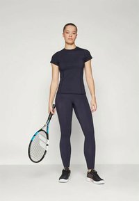 Mornarsko modra športna majica in legice, prilegajoča se zasnova, gladka tekstura, v paru s črnim in modrim teniškim rekvetom, stoji na enobarvni ozadju.