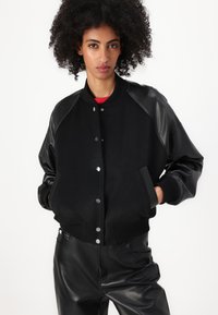 Čierna bomber bunda s vlnovým telom a koženými rukávmi. Obsahuje cvoky, rebrované manžety a dva bočné vrecká. Nosené s čiernymi nohavicami.