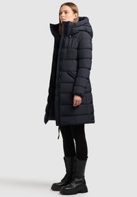 Námořnická modrá péřová bunda s kapucí, prošívaný design, zapínání na zip vpředu a dvě boční kapsy, spárována s černými legínami a černými šněrovacími botami.