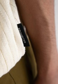 Cream-colored knit garment with textured stripes, featuring a black tag displaying the word "PROHIBITED." Background includes an olive green fabric.