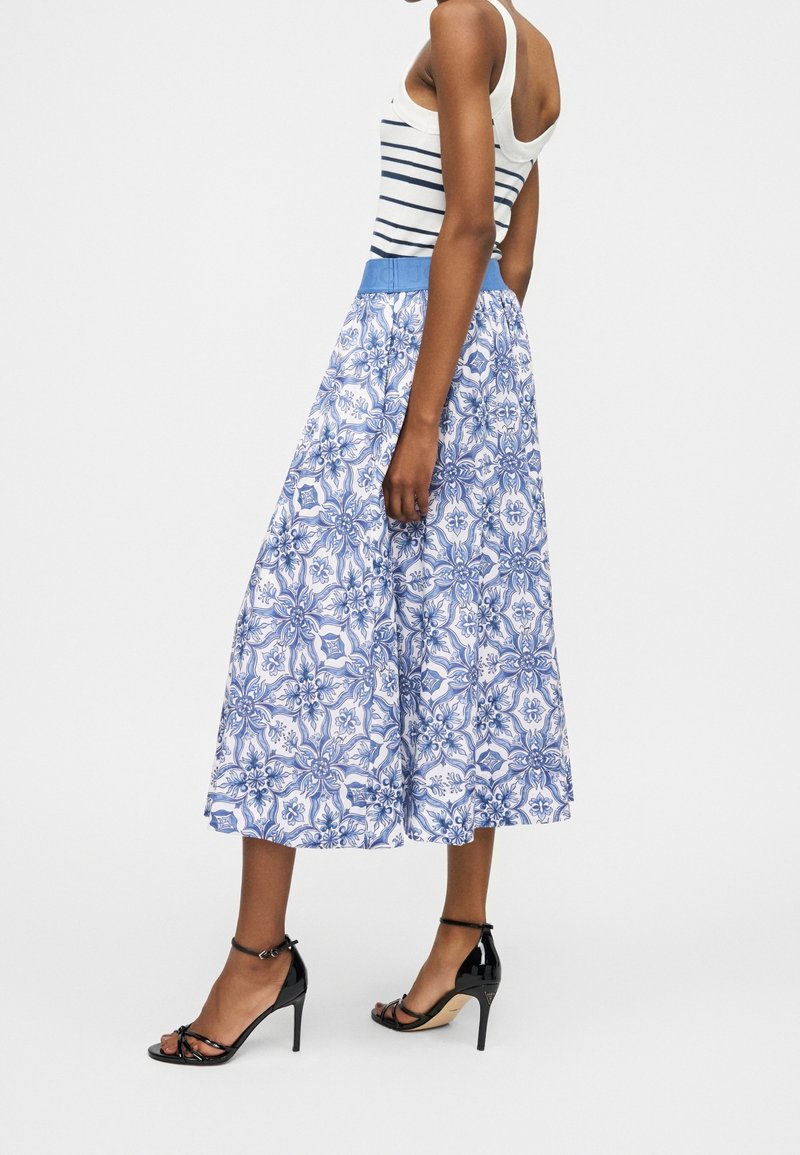 Femme portant un débardeur rayé blanc et bleu marine avec une jupe midi à motifs bleus et des sandales à talons noirs sur un fond uni.