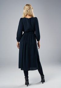 Kobieta z blond włosami ułożonymi w półwarkocz, ubrana w czarną midi sukienkę z długim rękawem, czarne legginsy i czarne wysokie botki za kostkę, stoi.