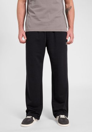 Man draagt zwarte losse broek, grijs shirt met korte mouwen en zwart-witte sneakers, staand tegen een effen achtergrond.