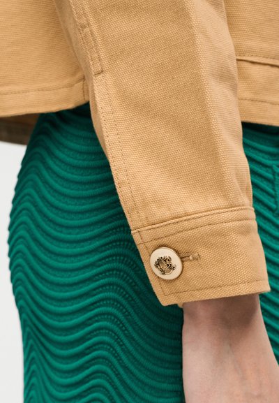 Μανίκι μπεζ σακακιού με διακοσμητικό κουμπί πάνω σε υφασμάτινη φούστα σε αποχρώσεις του τυρκουάζ, δείχνοντας λεπτομέρειες υφάσματος και ραφές στο ένδυμα.