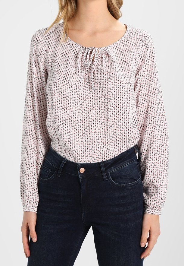 Helles rosa Langarmbluse mit gerafftem Ausschnitt, versehen mit einem sich wiederholenden kleinen Muster in Rot und Blau, kombiniert mit dunkelblauen Jeans.