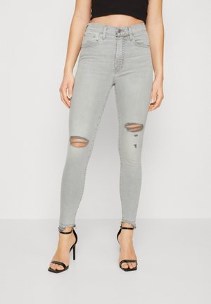Kvinde iført lysegrå slidte skinny jeans med frynsede kanter og sorte højhælede sandaler, stående mod en ensfarvet baggrund.