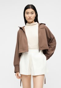 Νέα γυναίκα με ίσια μαύρα μαλλιά, που φοράει καφέ κοντή ζακέτα με φερμουάρ, μπεζ τοπ και λευκά σορτς με ψηλή μέση, στέκεται μπροστά σε λευκό φόντο.